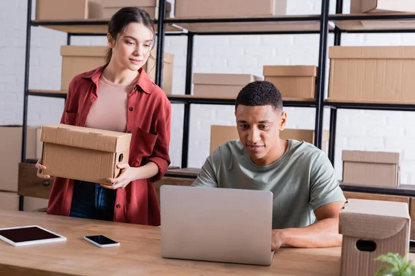 Vendedor celebración caja de cartón cerca de un colega afroamericano utilizando el ordenador portátil en la tienda web en línea - foto de stock