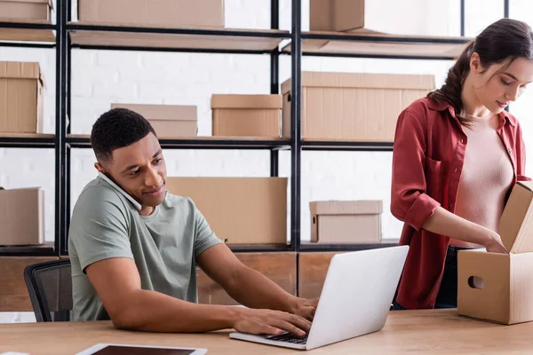 African american seller talking on smartphone and using laptop near colleague with box in online web store — Stock Photo