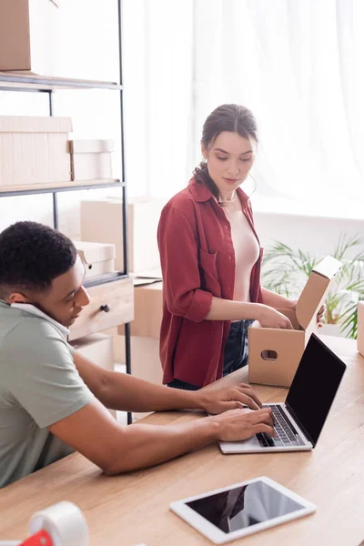 Scatola di imballaggio venditore vicino al collega afro-americano che utilizza laptop e parla su smartphone nel negozio online — Foto stock