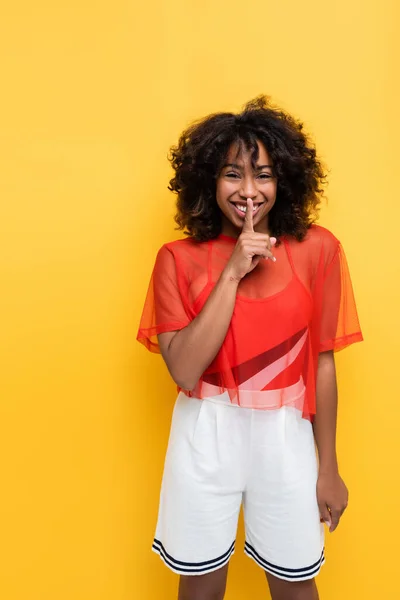 Cheerful african american woman in white shorts showing hush gesture isolated on yellow — Stock Photo