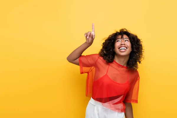 Sonriente mujer afroamericana en ropa de verano mirando hacia arriba y señalando con el dedo aislado en amarillo - foto de stock