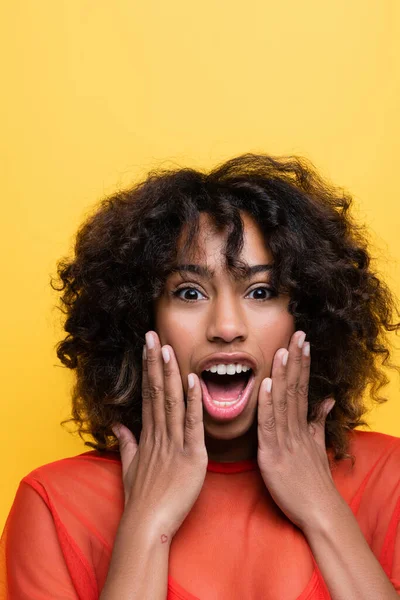 Amazed african american woman touching face and looking at camera isolated on yellow — Stock Photo