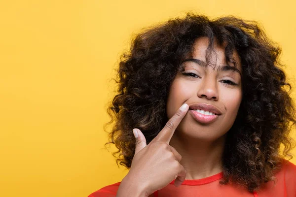 Alegre afroamericana mujer apuntando con el dedo a su propia sonrisa aislado en amarillo - foto de stock