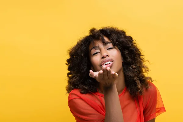 Bonita e sorridente afro-americana soprando beijo de ar isolado no amarelo — Fotografia de Stock