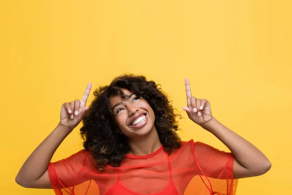 Femme afro-américaine souriante levant les yeux et pointant du doigt isolé sur jaune — Photo de stock