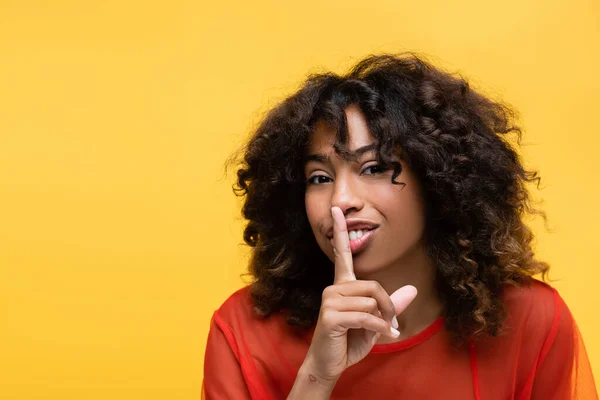 Sorrindo mulher afro-americana mostrando sinal de silêncio e olhando para a câmera isolada no amarelo — Fotografia de Stock