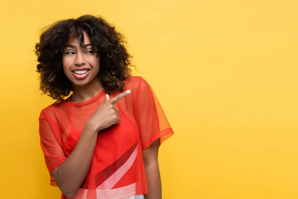 Happy african american woman in red top looking away and pointing with finger isolated on yellow — Stock Photo