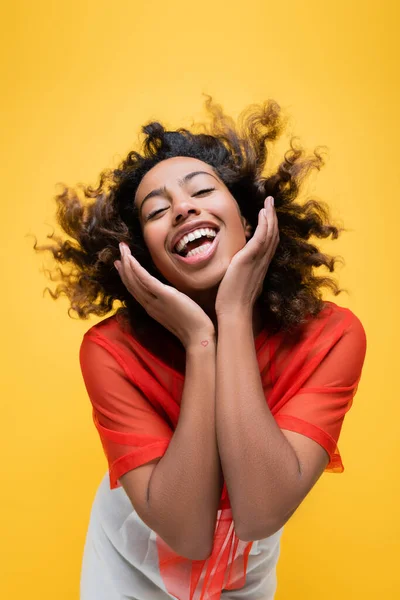 Здивована і щаслива афроамериканська жінка тримає руки біля обличчя ізольовано на жовтому — стокове фото