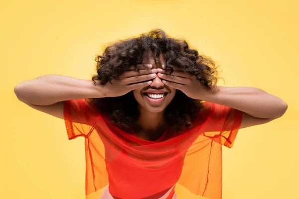 Mujer afro-americana feliz en verano cubriendo los ojos con las manos aisladas en amarillo - foto de stock
