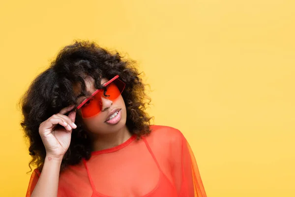Morena afroamericana americana mujer ajustando gafas de sol y mirando hacia otro lado aislado en amarillo - foto de stock