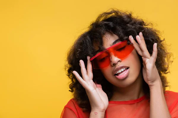 Чувственная африканская американка с волнистыми волосами регулирует модные солнцезащитные очки, изолированные на желтом — стоковое фото