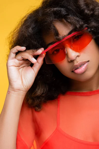 Mujer afroamericana de moda tocando gafas de sol rojas y mirando a la cámara aislada en amarillo - foto de stock