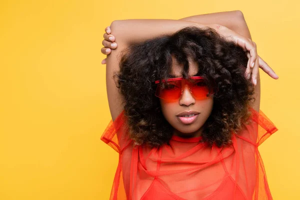 Femme afro-américaine en haut rouge et des lunettes de soleil à la mode posant avec les mains au-dessus de la tête isolé sur jaune — Photo de stock