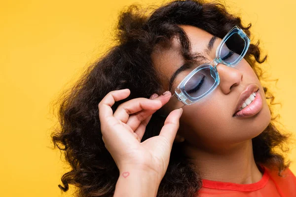 Mulher afro-americana com olhos fechados ajustando óculos azuis isolados no amarelo — Fotografia de Stock