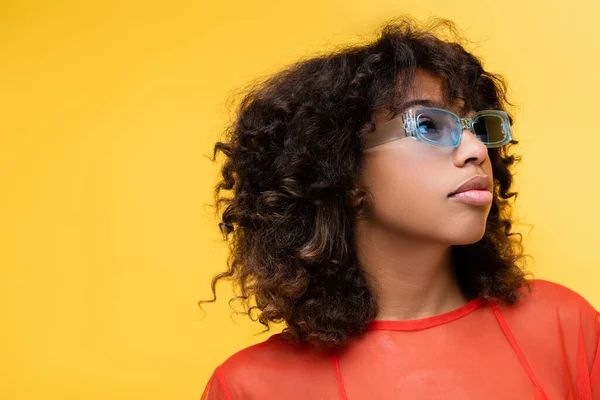 Encaracolado Africano americano mulher em azul óculos de sol elegantes olhando para longe isolado no amarelo — Fotografia de Stock