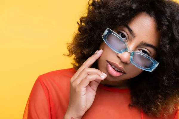 Sensual mulher americana africana em óculos de sol azuis tocando rosto e olhando para a câmera isolada no amarelo — Fotografia de Stock