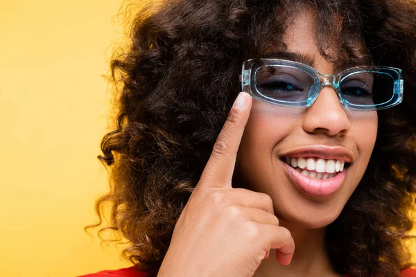 Усміхнена афроамериканка вказує на блакитні стильні сонцезахисні окуляри ізольовані на жовтому — стокове фото