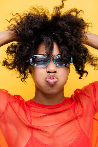 Mujer afroamericana en gafas de sol de moda haciendo pucheros labios y tocando el pelo ondulado aislado en amarillo - foto de stock