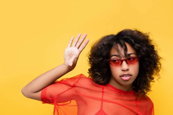 Модна афроамериканська жінка в червоному верху і сонцезахисні окуляри махають рукою ізольовано на жовтому — стокове фото