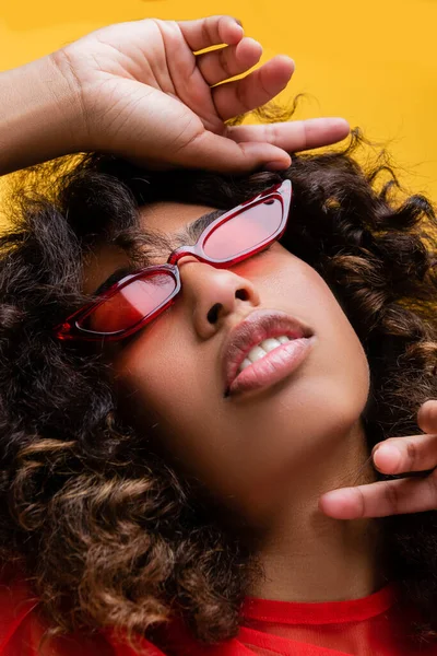 Primer plano retrato de la sensual mujer afroamericana en gafas de sol cogidas de la mano cerca de la cara aislada en amarillo - foto de stock