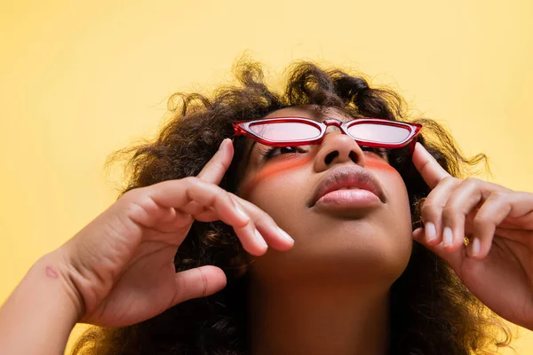 Vista basso angolo di donna afro-americana regolazione occhiali da sole alla moda e guardando in alto isolato sul giallo — Foto stock