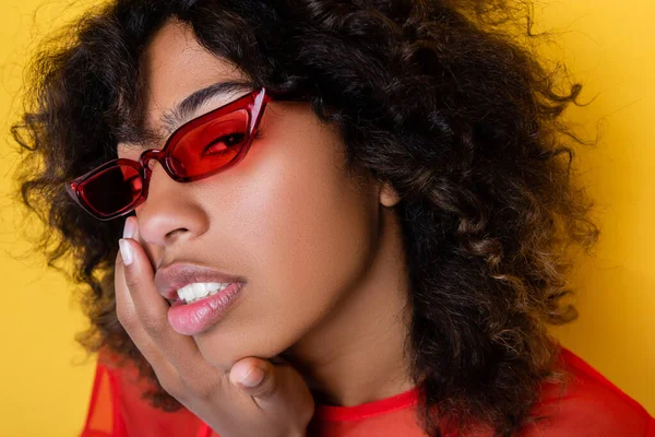 Porträt einer afrikanisch-amerikanischen Frau mit roter Sonnenbrille, die ihr Gesicht isoliert auf gelb berührt — Stockfoto