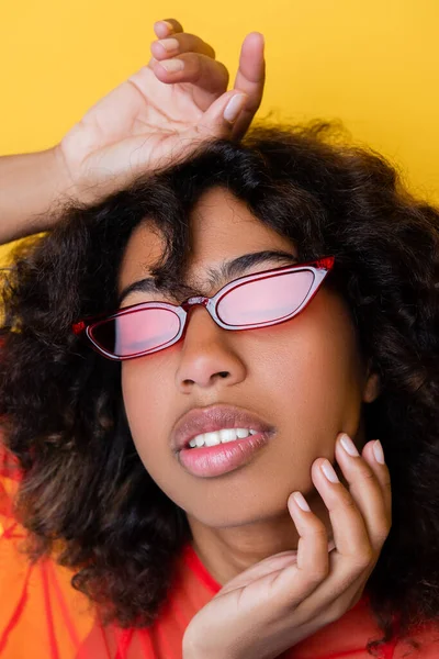 Portrait of curly african american woman in trendy sunglasses touching face isolated on yellow — Stock Photo