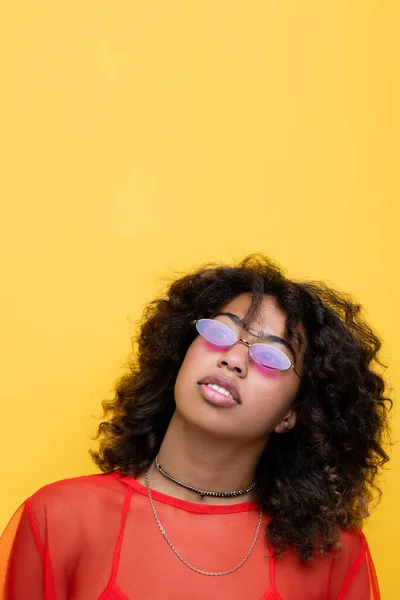 Morena afroamericana americana mujer en gafas de sol con estilo y collares mirando hacia arriba aislado en amarillo - foto de stock