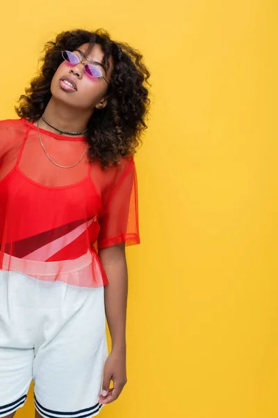 Femme afro-américaine en haut rouge, short blanc et lunettes de soleil regardant loin isolé sur jaune — Photo de stock