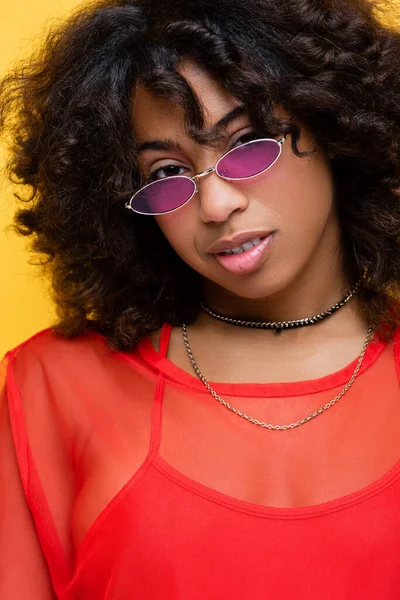 Bruna donna afroamericana in collane e occhiali da sole alla moda sorridenti alla fotocamera isolata sul giallo — Foto stock
