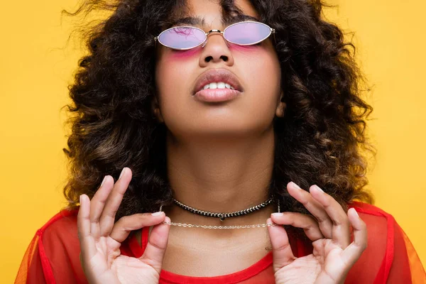 Кучерява афроамериканська жінка в модних сонцезахисних окулярах, що торкаються намистах ізольована на жовтому — стокове фото