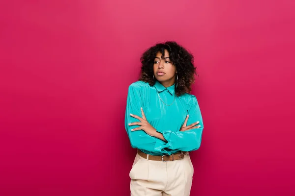 Mujer afroamericana con el pelo ondulado de pie con los brazos cruzados y mirando hacia otro lado aislado en rosa - foto de stock