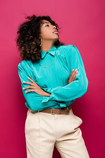 Mujer afroamericana en blusa azul de pie con los brazos cruzados y mirando hacia otro lado aislado en rosa - foto de stock