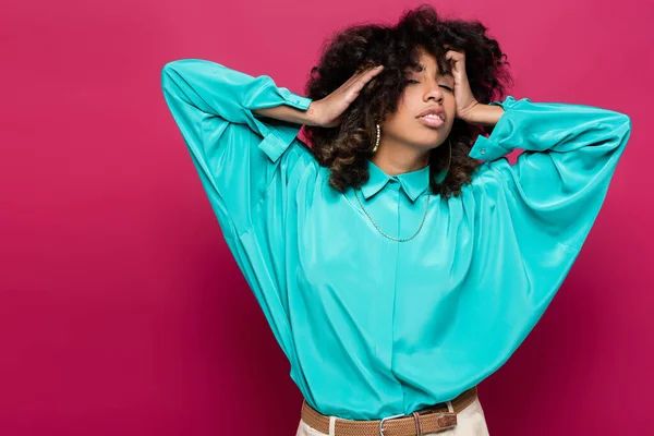 Mujer afroamericana de moda tocando el pelo ondulado mientras posa en blusa azul aislada en rosa - foto de stock