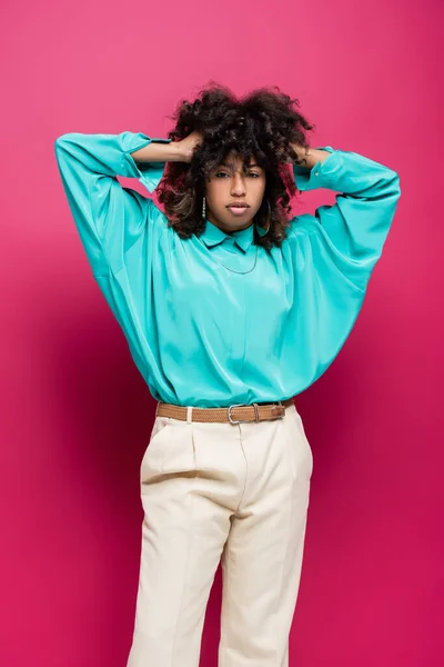 Trendy african american woman in turquoise blouse touching curly hair isolated on pink — Stock Photo