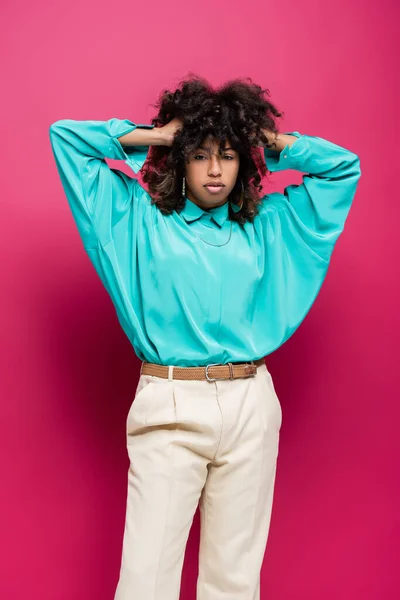 Mulher americana africana na moda em blusa turquesa tocando cabelo encaracolado isolado em rosa — Fotografia de Stock