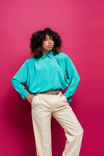 Femme afro-américaine en chemisier turquoise posant les mains dans des poches de pantalon isolé sur rose — Photo de stock