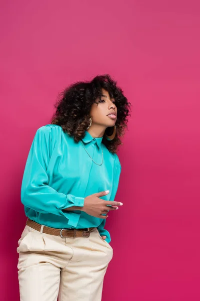 Femme afro-américaine en chemisier turquoise debout avec la main dans la poche et regardant loin isolé sur rose — Photo de stock