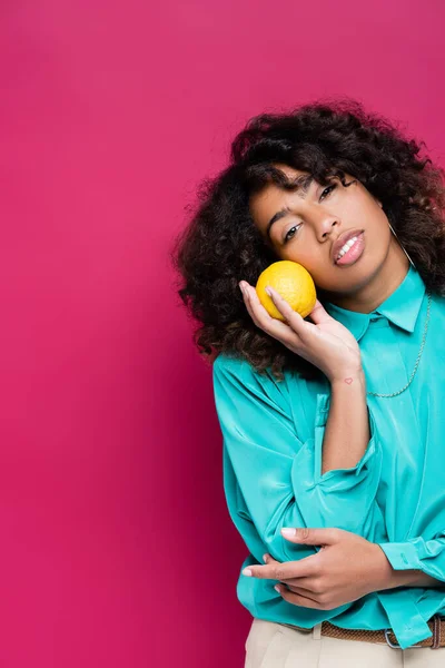 Donna afroamericana con i capelli ondulati tenendo arancio maturo isolato su rosa — Foto stock