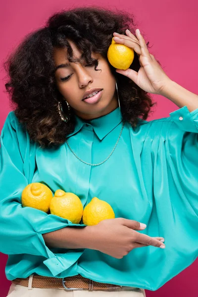 Morena mujer afroamericana en blusa turquesa posando con limones aislados en rosa - foto de stock