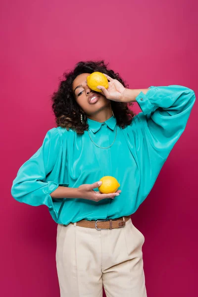 Mujer afroamericana en blusa azul oscureciendo la cara con limón aislado en rosa - foto de stock