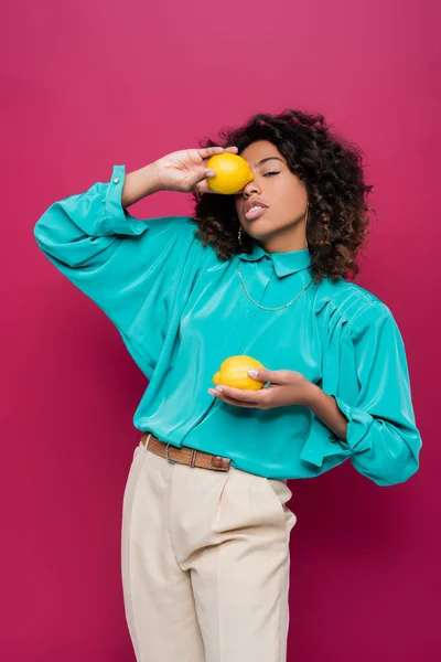 Mujer afroamericana en blusa turquesa oscureciendo la cara con limón entero aislado en rosa - foto de stock