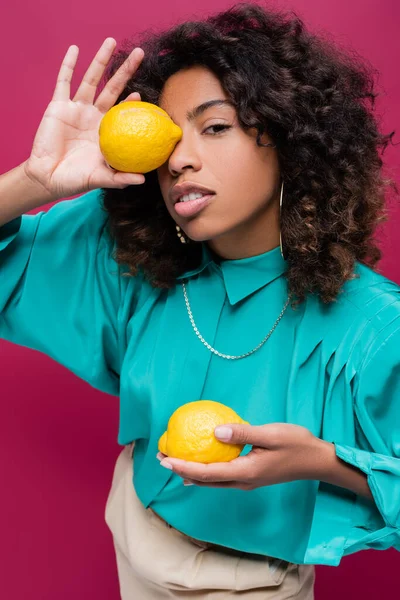 Pretty african american woman in cyan blouse obscuring face with lemon isolated on pink — Stock Photo