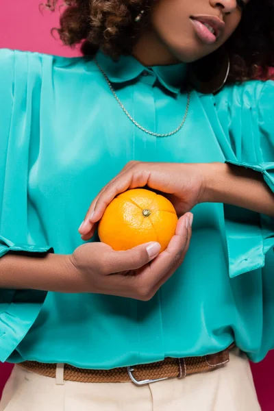 Частковий вид на афроамериканку в бірюзовій блузці, що тримає весь апельсин ізольований на рожевому — стокове фото