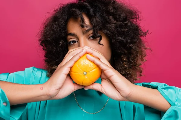 Curly african american woman obscuring face with ripe orange isolated on pink — Stock Photo