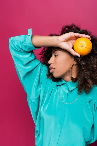 Curly african american woman posing with ripe orange isolated on pink — Stock Photo