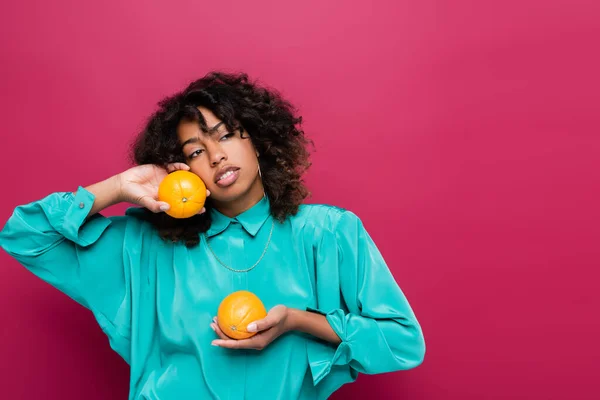 Африканская американка в бирюзовой блузке держит апельсины и смотрит в сторону изолированных на розовый — стоковое фото