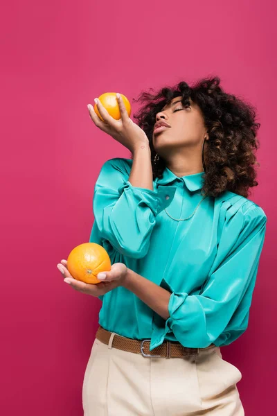 Кучерява афроамериканська жінка в стильній блузці позує з апельсинами ізольовано на рожевому — стокове фото