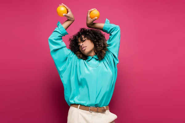 Femme afro-américaine en chemisier turquoise posant avec des oranges dans les mains levées isolé sur rose — Photo de stock