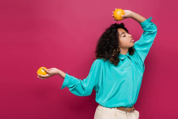 Femme afro-américaine à la mode posant les yeux fermés et des oranges mûres isolées sur rose — Photo de stock
